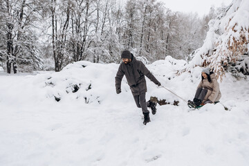 Young couple having fun sledging down pulling sled sleigh in winter snowy season in city park covered hill, holiday vacation weekend, enjoying spending time together
