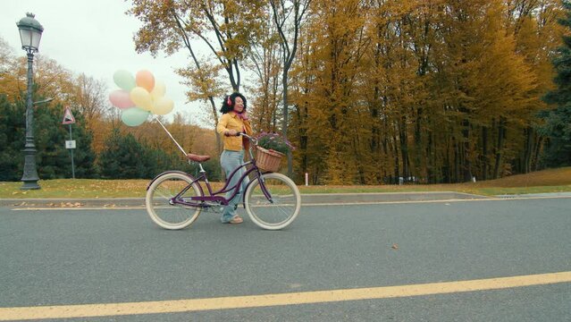 Curly dark-haired female in red headphones strolling on a fine autumn weather with a ladies bicycle decorated with balloons and basket with flowers. High quality 4k footage