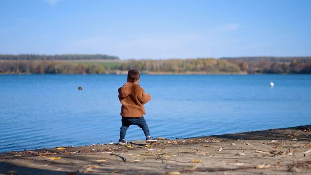 Baby boy spending sunny autumn day at the river. Caucasian toddler throwing the stones into water.