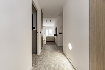 Distributor hallway of a modern house with gray terrazzo floor, smooth white painted walls, access to several rooms and light integrated into the wall