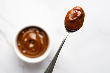 A spoonful and a bowl full of dulce de leche on white marble background with copy space. 