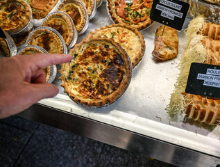 Paris, France. June 30 2022. At the beautiful Marché Popincourt local market, a food stall with French savory pie specialities. A customer's hand indicates which one to buy. - obrazy, fototapety, plakaty