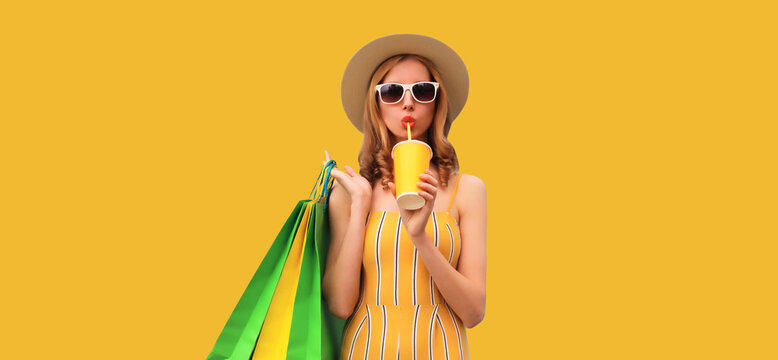 Beautiful stylish young woman posing with shopping bags and drinks fresh juice wearing summer straw hat on yellow studio background