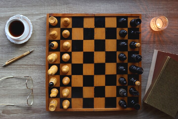 Vintage wooden chessboard, books, glasses, pen, cup of tea or coffee and scented candle on the table. Dark academia concept. Top view.