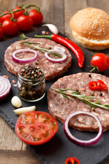 Raw beef hamburger patties on stone plate, vegetables and spices, wooden background.