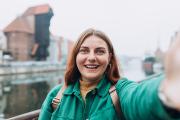 Young happy 30s woman is making selfie on a camera on city street in Gdansk. Urban life concept. Girl talking on a video call with phone outdoor. Traveling Europe in autumn. Famous Zuraw crane