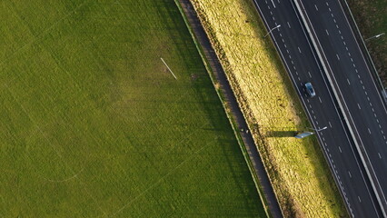 Overhead aerial photography of a public football field and a highway separated by a cycle path with cyclist and a car passing - obrazy, fototapety, plakaty
