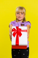 Fototapeta premium Lovely smiling preteen child girl kid presenting birthday gift box stretches out hands, offer wrapped present career bonus, celebrating party. Little children on studio yellow background. Vertical