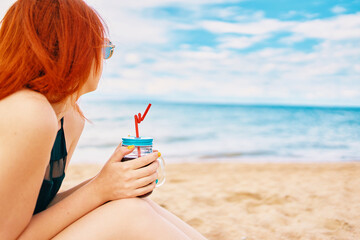 Cute girl in a swimsuit is sitting on beach with a cocktail and look away. Tropical alcohol beverage. Summer day with cloudy sky. Red-haired woman with sunglasses on vacation. Holidays at the sea.