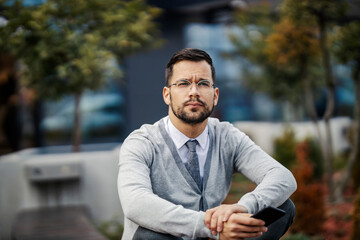 Fototapeta premium A young elegant man is sitting with legs crossed on a bench and holding phone.