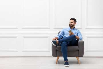 Handsome man with smartphone sitting in armchair near white wall indoors, space for text
