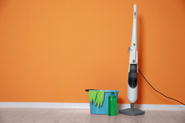 Modern steam mop, bucket with gloves and bottle of cleaning product on floor near orange wall,...