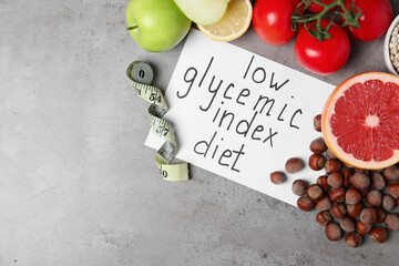 Paper with words Low Glycemic Index Diet, measuring tape and food on grey table, flat lay. Space for text
