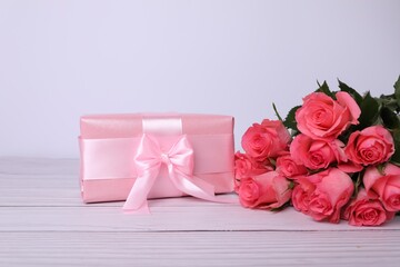 Gift box with pink bow and bouquet of beautiful roses on white wooden table, space for text