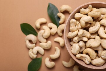 Tasty cashew nuts and green leaves on pale brown background, top view