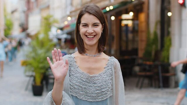 Hello. Young pretty woman smiling friendly at camera, waving hands gesturing hi, greeting or goodbye, welcoming with hospitable expression outdoors. Happy lovely girl walking in urban city town street