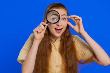 Investigator researcher scientist woman holding magnifying glass near face, looking into camera with big zoomed funny eye, searching, analysing. Pretty girl isolated on blue studio background indoors