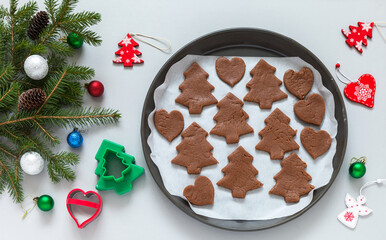Homemade Christmas cookies in shape of trees and hearts made from chocolate dough in baking dish. Cookie cutters, Christmas tree branches with toys on light table. New Year and Christmas chores