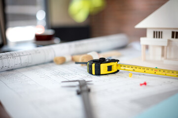 Tape measure placed with architectural equipment, tools, yellow safety helmet, house model and blueprint scatter around on meeting table for residential building and renovation project at office