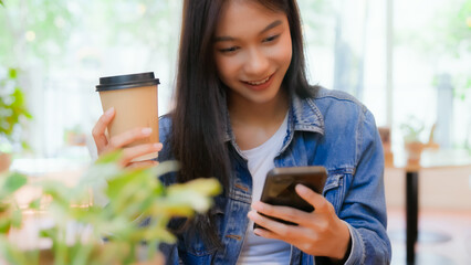 Asian woman happy and typing messages on smartphone in cafe. Smiling young woman sitting at table...