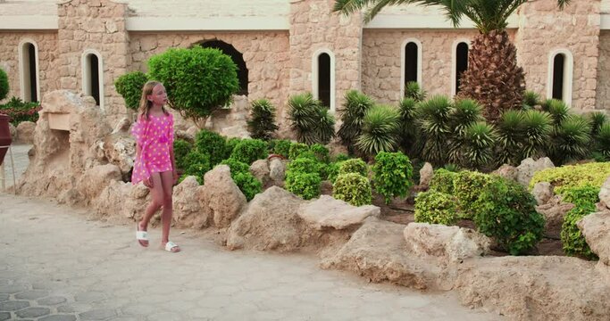 Teenage girl walking by rocks and plants at park. Handheld shot of female is spending leisure time at luxurious hotel. She is enjoying her vacation in historic place.