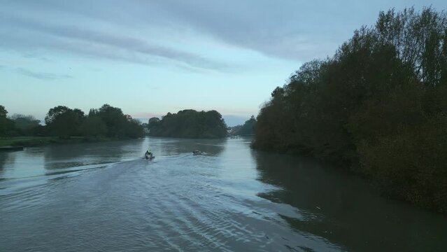 Rowing boat sculling on River Thames Richmond in grey light of dawn. A coach's motorboat travels behind it. Autumnal Trees.