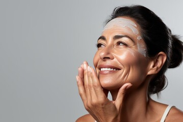 Close-up of middle-aged Caucasian woman touching her face to apply moisturizer. Smiling face of adult brunette lady with daily cream, facial cosmetics. Skin care. Grey background, copy space.