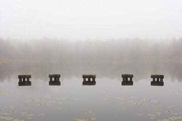 A beautiful foggy morning on the shore of a pond. Reflections of trees and rocks in the water. Foggy autumn landscape. Calm European landscape. The remains of the old bridge.