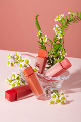 Four unbranded lipsticks are displayed alongside daisies and fern leaves on a pink background. Concept of homemade cosmetics with natural ingredients.
