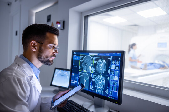Doctor examining X-ray images on display in MRI control room while in background nurse preparing the patient for examination test.