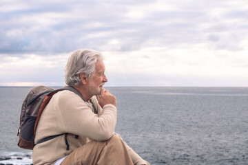 Fototapeta na wymiar Adult senior man sitting on the cliff in sea excursion looking at horizon over water. Caucasian white haired grandfather holding backpack enjoying relax and retirement.
