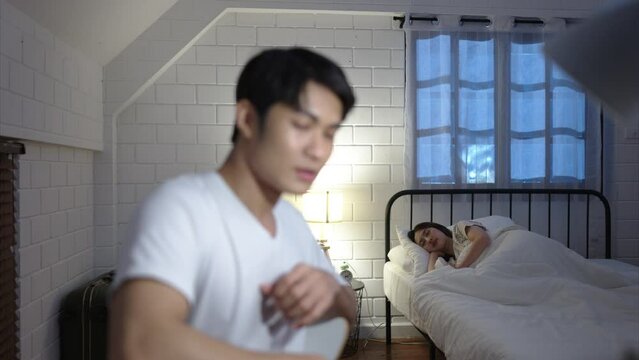 A young Asian man working overtime at home glance at his wife who was sleeping on the bed. prior to continuing work