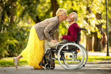 Senior couple in love. Cute photo of elderly woman kissing her husband in wheelchair. - obrazy, fototapety, plakaty