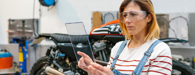 Concentrated female mechanic with security glasses holding transparent tablet in front of...
