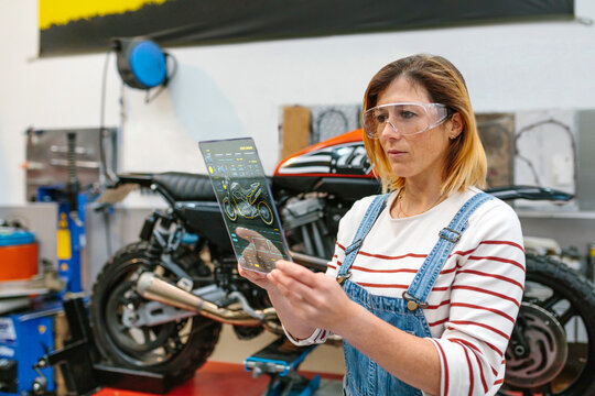 Concentrated female mechanic with security glasses touching transparent digital tablet with diagnosis app to review motorcycle on factory