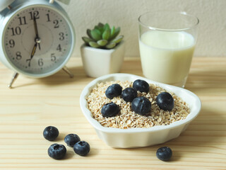 healthy breakfast with oat or granola in heart shape bowl, fresh blueberries, milk and white alarm clock 7 am on wooden table. Healthy breakfast concept.