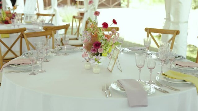 Close-up bouquet of wild flowers in pink colors are on the festive table. Minimalistic decor at the wedding, rounded table with white tablecloth, wine glasses and plates on the table. slow motion.