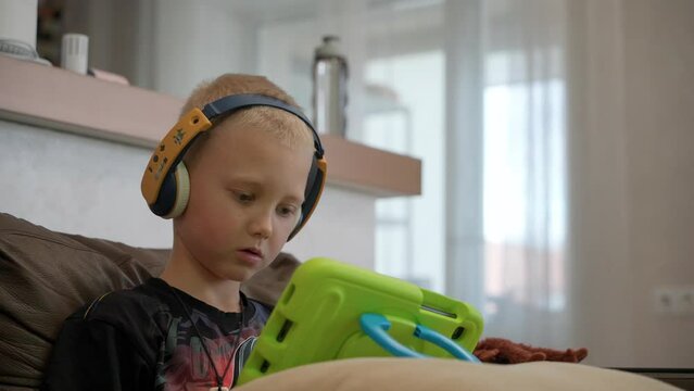 A child wearing headphones plays games on a tablet while sitting on the couch at home. A boy with blond hair using a digital tablet to watch cartoons or play online games.