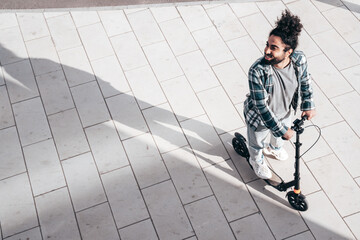 Trendy smiling bearded man in casual clothes riding electric scooter on a road in urban background. Handsome model posing in the street. Hipster guy with curly hairstyle. Top view