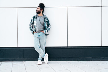 Handsome smiling hipster model. Unshaven Arabian man dressed in summer casual clothes, jeans and shirt. Fashion male with long curly hairstyle posing in the street. Near urban wall