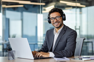 Portrait of successful businessman inside office, man at workplace smiling and looking at camera, using headphones and laptop at work, Indian programmer in business suit.