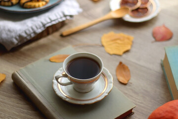 Cup of tea or coffee, plate with desserts, dried oranges, bowl of grapes, scented candles, vintage books, pumpkins and autumn leaves on the table. Autumnal hygge. Selective focus.