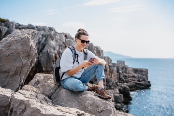 Woman traveler hiker relaxing on a rocky seashore with a smartphone in her hands. Summer travel and adventure concept