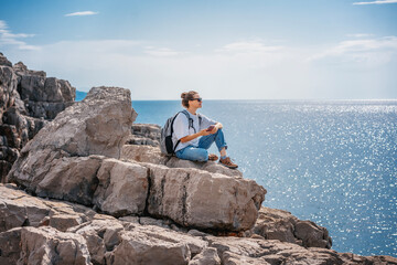 Woman traveler hiker relaxing on a rocky seashore with a smartphone in her hands. Summer travel and...