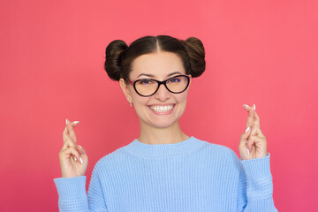 a young pretty happy woman in blue clothes and glasses smiles and looks at the camera, fingers crossed for good luck. Isolated on a pink background.