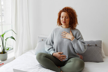 Portrait of plus-size curly redhead female in casual clothes doing pranayama technique sitting on...