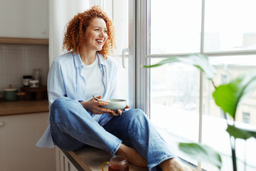 Portrait of happy young redhead female in jeans enjoying picturesque view from window during...