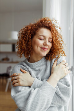 Gorgeous lovely charming redhead female with cute curls standing with closed eyes next to window at home, hugging herself, touching her favorite soft blue sweater, enjoying comfort of fabric