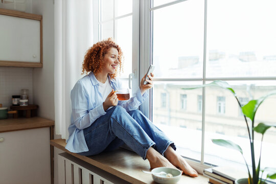 Woman using smartphone sitting on windowsill during breakfast holding cup of tea in hands, checking social media news feed, chatting in dating app, shopping online using marketplace applications