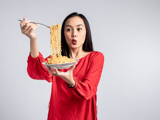 A portrait of a happy Asian woman wearing a red shirt, posing eating noodles. Isolated against a...